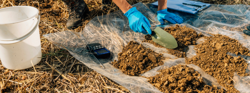Agronomist taking soil samples in a field with a bucket, soil-analyzer device, trowel, and clipboard